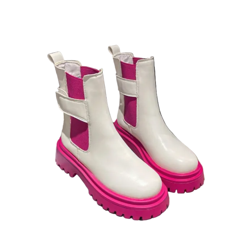 A pair of white Chelsea-style boots with bright pink elastic side panels and chunky pink soles, displayed on a beige surface with reflections in the background.