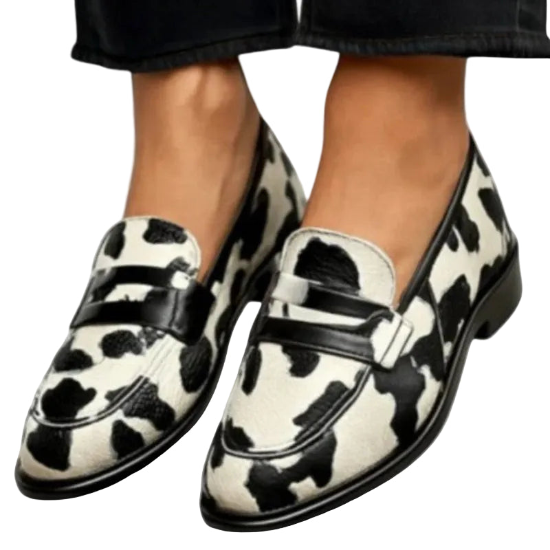 A pair of women's slip-on loafers with a black and off-white cow print pattern and black leather straps, shown on a model wearing black jeans. A small inset image in the top-left corner shows the shoes from a side angle.