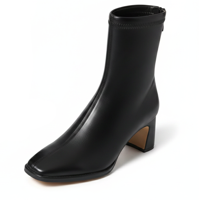 A pair of black leather ankle boots with pointed toes and block heels, displayed on crumpled white tissue paper with a black shoebox in the background.