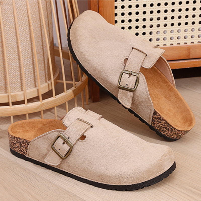 Pair of beige suede clogs with buckles on a wooden floor.